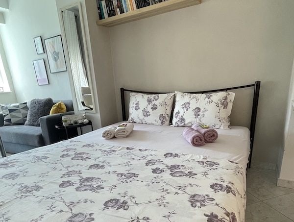 Cozy apartment interior with a neatly made double bed dressed in romantic and modern bedding, complemented by a stylish gray sofa adorned with beautiful decorative pillows in the background.